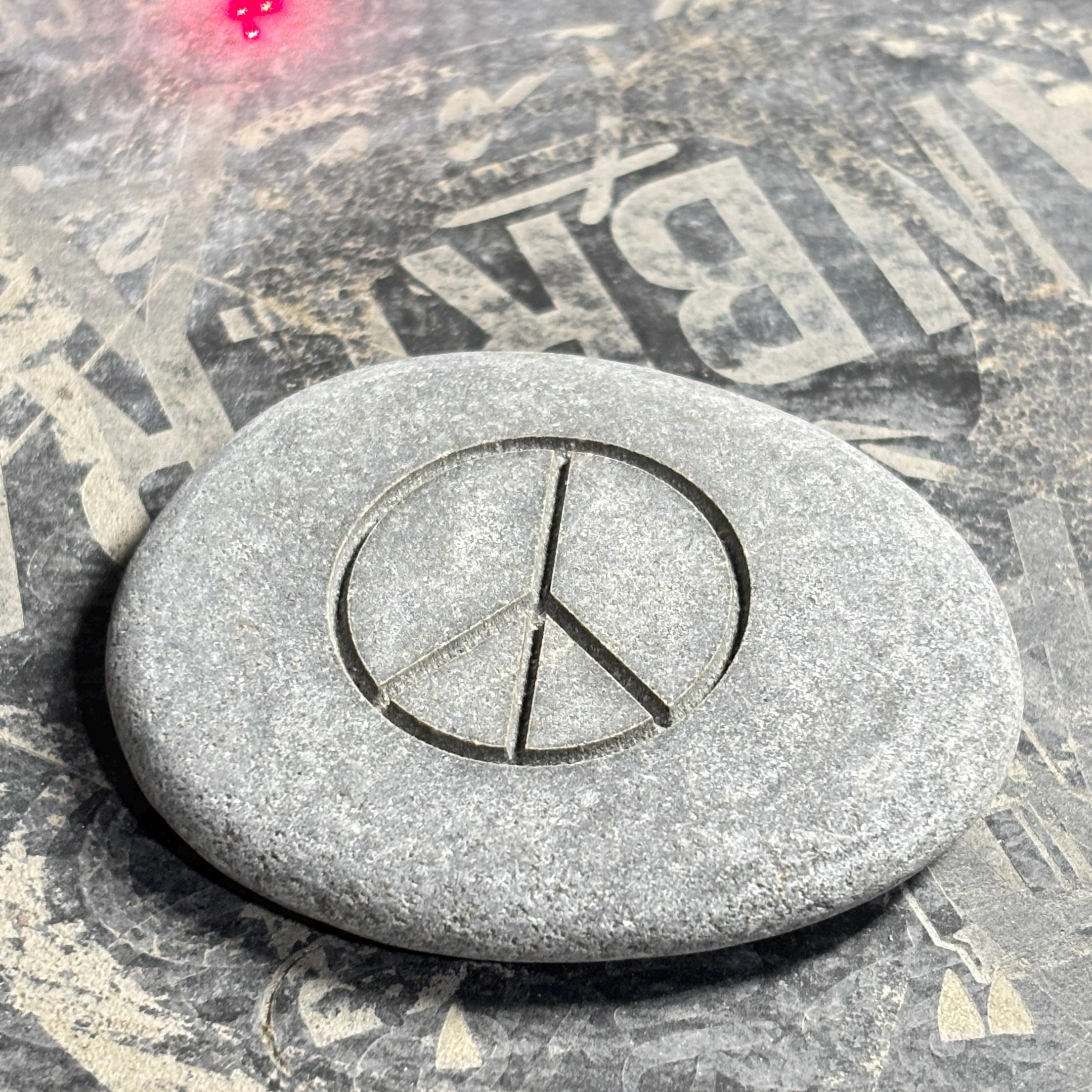 A grey river stone engraved with a peace symbol rests on a surface with bold lettering.