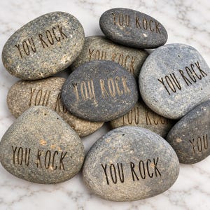 May include: A collection of smooth, grey river stones, each engraved with the text "YOU ROCK" in a simple, sans-serif font. The stones vary in size and shape, featuring natural speckled textures and shades of charcoal, slate, and light grey.