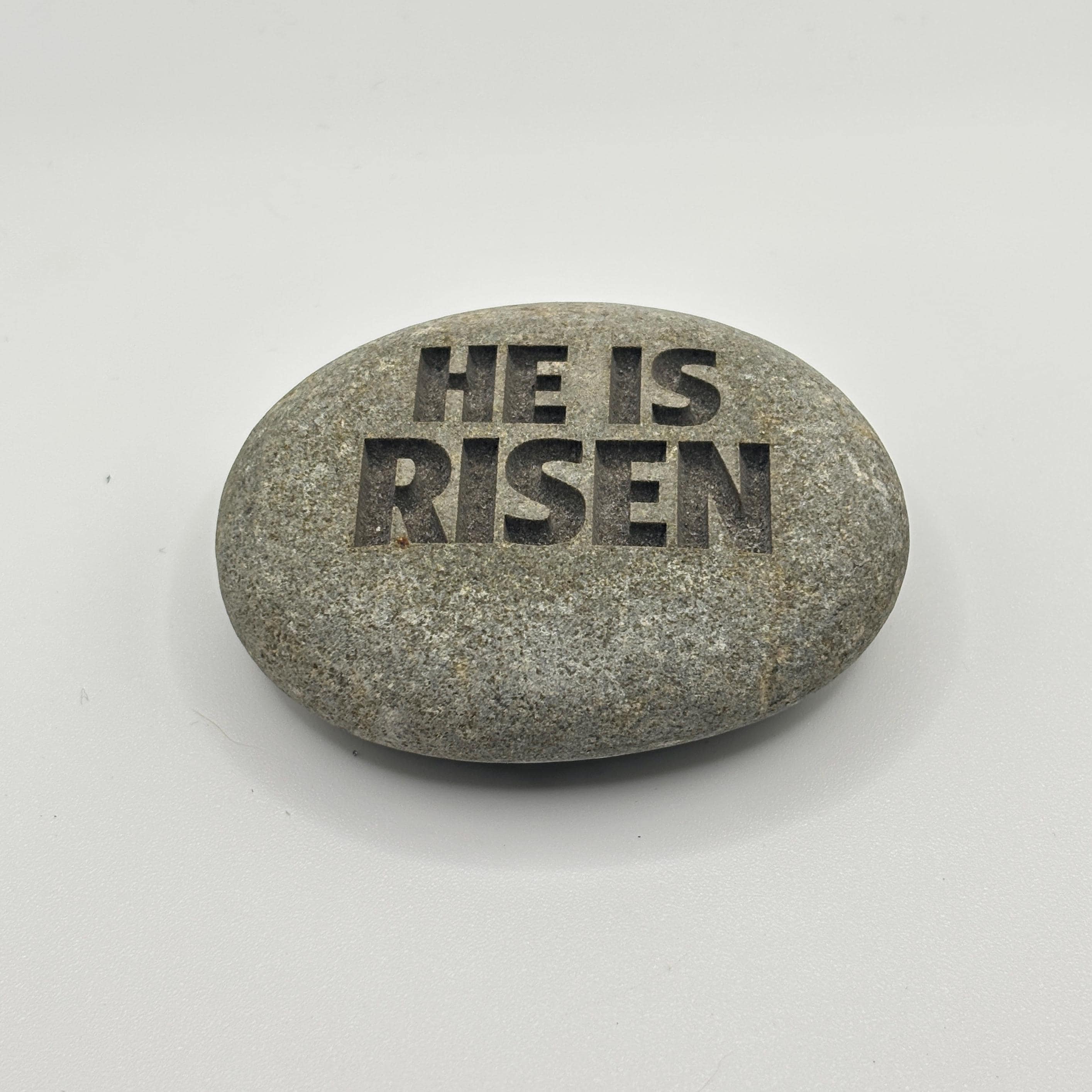A grey river stone with the engraved words "HE IS RISEN".