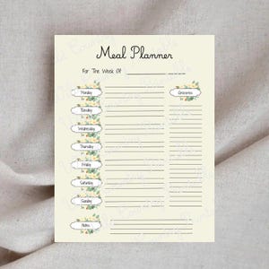 Puede incluir: Un planificador de comidas color crema con detalles florales. En la parte superior se lee "Meal Planner" y "For The Week Of". Incluye secciones para cada día de la semana, compras y notas. El diseño presenta delicados adornos florales.