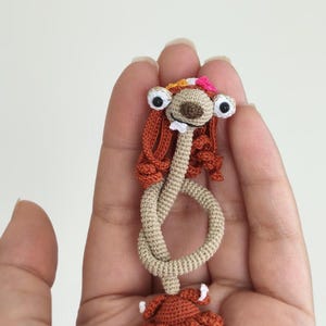 May include: A handmade crochet doll with large googly eyes, a floral headband, and rust-colored hair and dress. The doll's body is a light beige color, and the arms are knotted. The doll is held in a hand.