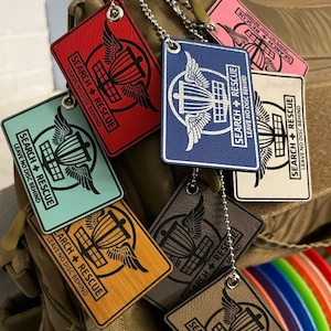 Könnte beinhalten: Ein Satz von sieben Disc-Golf-Taschenanhängern mit einem Disc-Golf-Korb und Flügeln. Die Anhänger sind aus verschiedenen Materialien gefertigt, darunter Holz, Leder und Metall. Jeder Anhänger trägt den Text "Search + Rescue" und "Leave No Disc Behind".