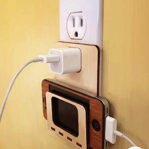 May include: A light-colored wooden wall-mounted phone charging dock featuring a rectangular cutout for a smartphone, a slot for a power adapter, and a white wall outlet plate. The dock is designed to hold a device horizontally while charging.