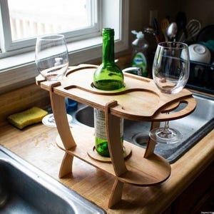 Puede incluir: Un soporte de vino de madera con una botella de vino verde y dos copas de vino. El soporte es de madera clara y tiene recortes para la botella y las copas. Las copas son transparentes.