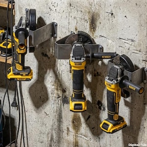 Puede incluir: Tres amoladoras angulares DeWalt amarillas y negras montadas en una pared de hormigón. Cada amoladora está asegurada en un soporte metálico personalizado. Las amoladoras tienen mangos negros y están en un taller.