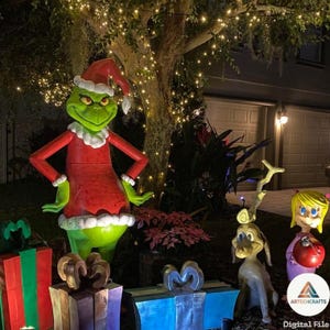 May include: A large green character wearing a red Santa suit and hat stands next to a brown dog with a single antler, a small blonde figure holding a red ornament, and several large gift boxes with bows in a nighttime outdoor display.