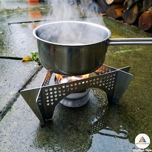 Può includere: Una pentola in acciaio inossidabile con manico è su un fornello da campeggio portatile, con vapore che sale dall'acqua bollente. Il fornello ha un telaio in metallo argentato con un design perforato. La scena è all'aperto.