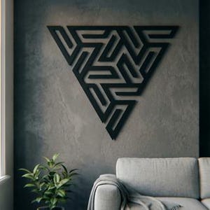 Könnte beinhalten: Ein schwarzes geometrisches Wandkunstwerk in Form eines Dreiecks, montiert an einer grau strukturierten Wand. Das Kunstwerk weist ein kompliziertes, sich wiederholendes Muster auf. Der Raum umfasst ein Sofa, Pflanzen und ein Fenster.