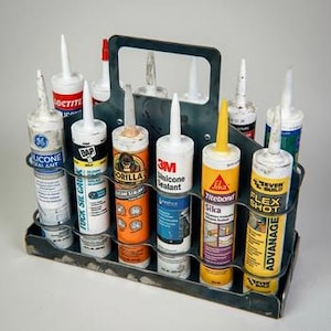 May include: A metal tool carrier filled with various tubes of construction sealant. The tubes are different colors and brands, including Loctite, DAP, Gorilla, 3M, and Titebond. The carrier has a handle and is sitting on a light gray surface.