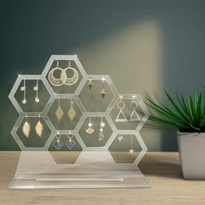 Op de afbeelding: Een heldere acryl oorbellen displaystandaard in een honingraatontwerp. De standaard bevat verschillende oorbellen, waaronder gouden hoepels, geometrische vormen en bungelende stijlen. De standaard staat op een houten oppervlak.