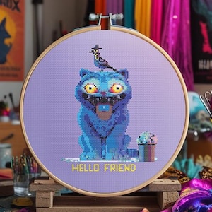 May include: A cross-stitch embroidery hoop featuring a blue cat with yellow eyes and a bird on its head. The text "HELLO FRIEND" is below the cat. The background includes art supplies and colorful fabric.