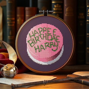 Può includere: Un telaio per punto croce con un disegno di torta rosa con le parole "Happy Birthday Harry" in verde. Il ricamo è ambientato su uno sfondo viola, incorniciato da un cerchio di legno. Il tema è una festa di compleanno.