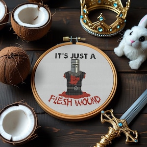 Puede incluir: Un bastidor de bordado de punto de cruz con el texto "IT'S JUST A FLESH WOUND" y un diseño de caballero. La imagen incluye cocos, un conejito de juguete, una corona dorada y una espada decorativa, todo dispuesto sobre una superficie de madera oscura.