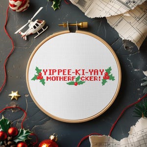 May include: A cross-stitch embroidery hoop with the text "YIPPEE-KI-YAY MOTHERFUCKER!" in red and green. The design includes holly leaves and berries. The hoop is set against a dark background with Christmas decorations.