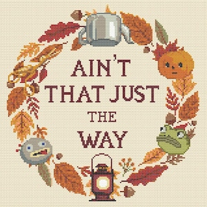 May include: Cross-stitch design featuring a circular wreath of autumn leaves in shades of orange, brown, and gold. The text "AIN'T THAT JUST THE WAY" is centered within the wreath. Includes cartoon images of a teapot, pumpkin, and lantern.