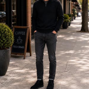 May include: A person wearing a black zip-up sweater, dark gray jeans, and black suede boots stands on a sidewalk. A sign for "Coffee" and "Fresh Brewed" is visible in the background.