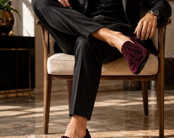Op de afbeelding: Bordeaux fluwelen loafers met een sierlijk patroon. De schoenen worden gecombineerd met een zwarte broek en een zwarte blazer. De loafers hebben een lage hak en een ronde neus. De afbeelding toont een persoon die in een houten stoel zit.