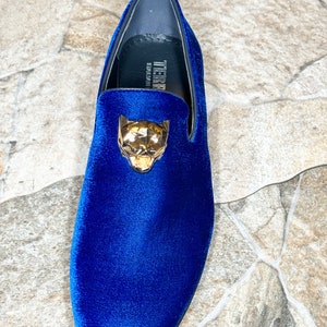 May include: A single blue velvet loafer with a gold-colored panther head ornament. The shoe has a rounded toe and a low heel. The brand name "TIERRA" is visible on the insole.