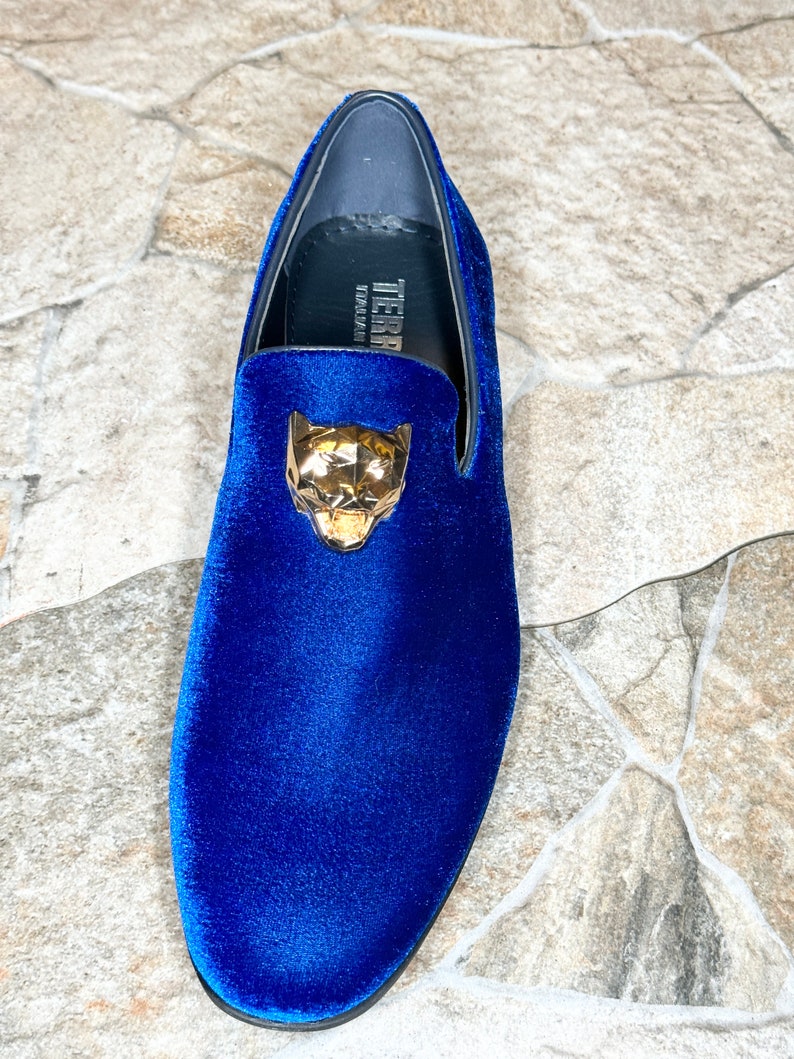 May include: A single blue velvet loafer with a gold-colored panther head ornament. The shoe has a rounded toe and a low heel. The brand name "TIERRA" is visible on the insole.