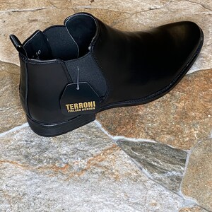 Puede incluir: Bot&iacute;n Chelsea de cuero negro con punta afilada. La bota tiene paneles laterales el&aacute;sticos y una leng&uuml;eta. Una etiqueta dice "TERRONI ITALIAN DESIGN". La bota se exhibe sobre una superficie de baldosas de piedra.