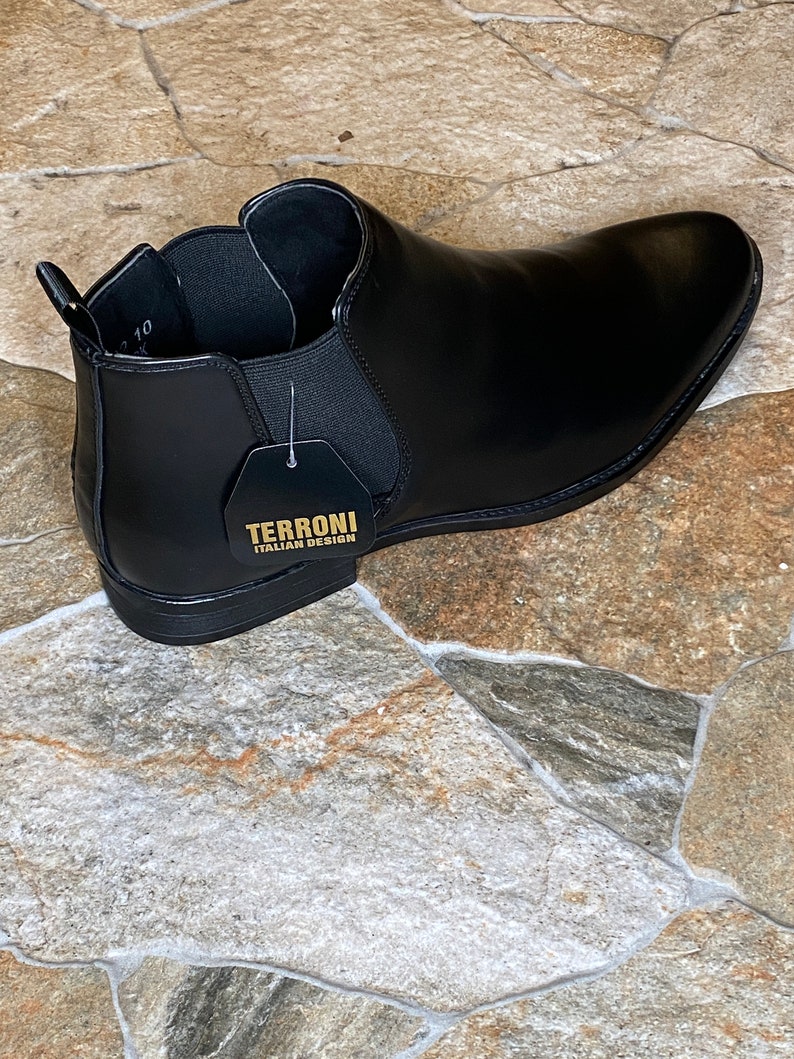 Puede incluir: Bot&iacute;n Chelsea de cuero negro con punta afilada. La bota tiene paneles laterales el&aacute;sticos y una leng&uuml;eta. Una etiqueta dice "TERRONI ITALIAN DESIGN". La bota se exhibe sobre una superficie de baldosas de piedra.