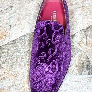Op de afbeelding: Een close-up van een enkele paarse fluwelen loafer met een sierlijk, verhoogd bloemmotief. De schoen heeft een ronde neus en een zwarte zool. De binnenkant is roze met de merknaam "TERRO DESIGNS" in het wit gedrukt.