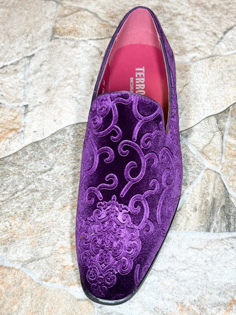 Op de afbeelding: Een close-up van een enkele paarse fluwelen loafer met een sierlijk, verhoogd bloemmotief. De schoen heeft een ronde neus en een zwarte zool. De binnenkant is roze met de merknaam "TERRO DESIGNS" in het wit gedrukt.