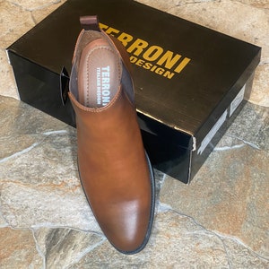 May include: A brown leather Chelsea boot with a pointed toe sits next to a black box. The box has gold lettering that reads "TERRONI DESIGN". The boot has a pull tab and elastic side panels. The insole reads "TERRONI ITALIAN DESIGN".