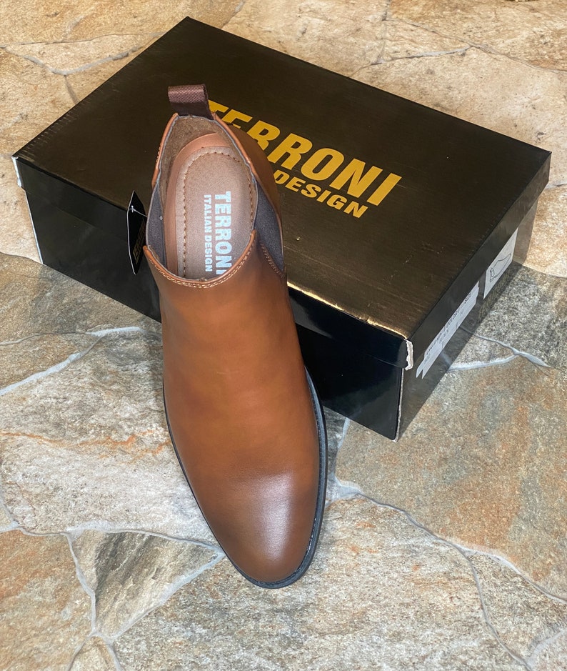 May include: A brown leather Chelsea boot with a pointed toe sits next to a black box. The box has gold lettering that reads "TERRONI DESIGN". The boot has a pull tab and elastic side panels. The insole reads "TERRONI ITALIAN DESIGN".