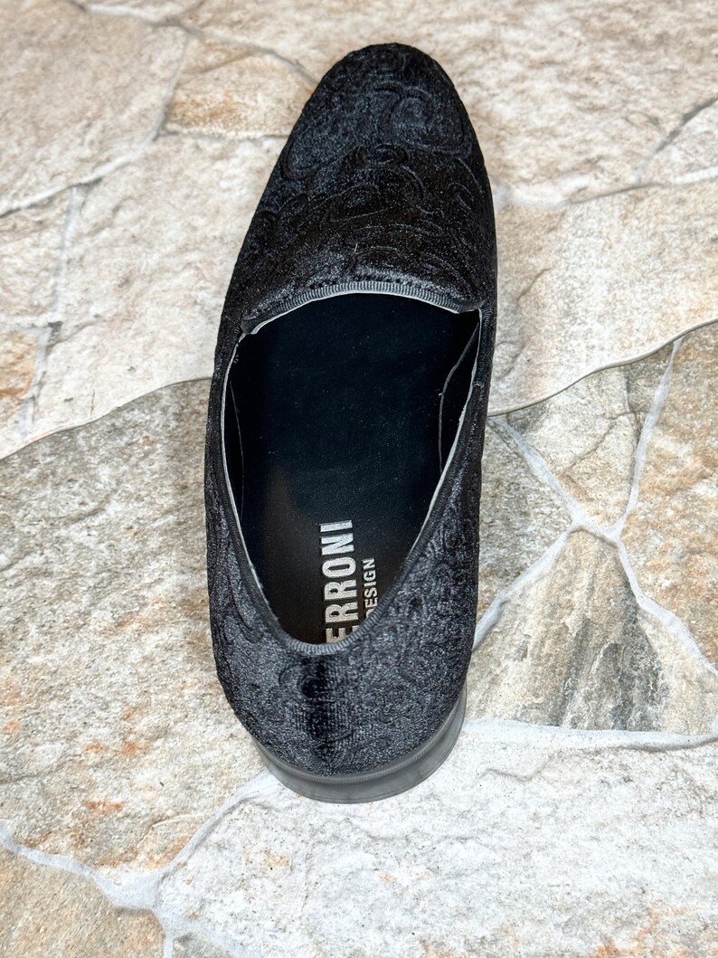 Puede incluir: Mocas&iacute;n negro de terciopelo con un estampado floral en relieve. El zapato tiene una puntera redondeada y un tac&oacute;n bajo. La marca "ERRONI DESIGN" es visible en la plantilla. Adecuado para ocasiones formales.