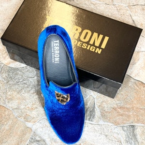 May include: A pair of royal blue velvet loafers with a gold-colored emblem. The shoes are displayed with a black box that has gold lettering that reads "TERRONI DESIGN". The shoes are a slip-on style.