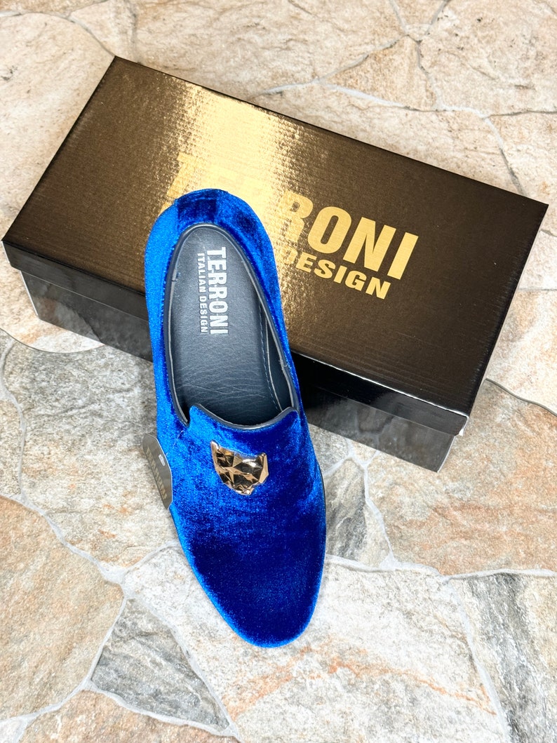 May include: A pair of royal blue velvet loafers with a gold-colored emblem. The shoes are displayed with a black box that has gold lettering that reads "TERRONI DESIGN". The shoes are a slip-on style.