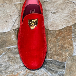 May include: A red velvet loafer with a gold-colored panther head emblem. The shoe has a rounded toe and a black sole. The interior is pink and burgundy. The shoe is a size 10.