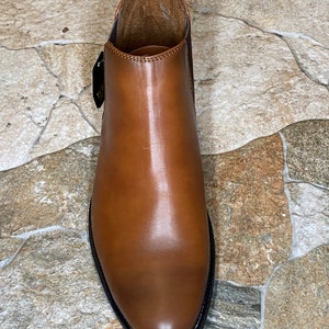 May include: A brown leather Chelsea boot with a rounded toe and a black sole. The boot has a pull tab at the back and a tag attached to the side. The leather has a polished finish.
