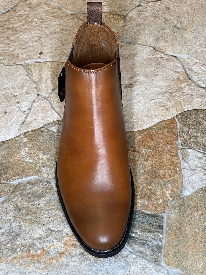 May include: A brown leather Chelsea boot with a rounded toe and a black sole. The boot has a pull tab at the back and a tag attached to the side. The leather has a polished finish.