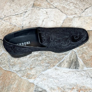 Puede incluir: Mocas&iacute;n de terciopelo negro con un intrincado estampado floral. El zapato tiene un tac&oacute;n bajo y una puntera redondeada. El nombre de la marca, "FERRONI", es visible en la plantilla. El zapato est&aacute; sobre un fondo de baldosas de piedra.