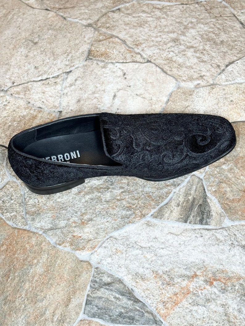 Puede incluir: Mocas&iacute;n de terciopelo negro con un intrincado estampado floral. El zapato tiene un tac&oacute;n bajo y una puntera redondeada. El nombre de la marca, "FERRONI", es visible en la plantilla. El zapato est&aacute; sobre un fondo de baldosas de piedra.