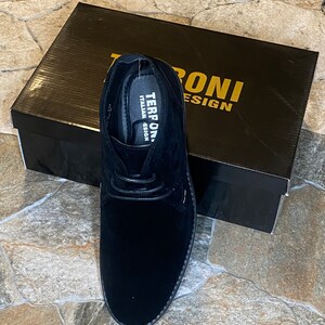 May include: Black suede chukka boot with a lace-up design, displayed next to a black box. The box features gold lettering that reads "TERRONI DESIGN". The shoe has a stitched sole and a soft, textured appearance.