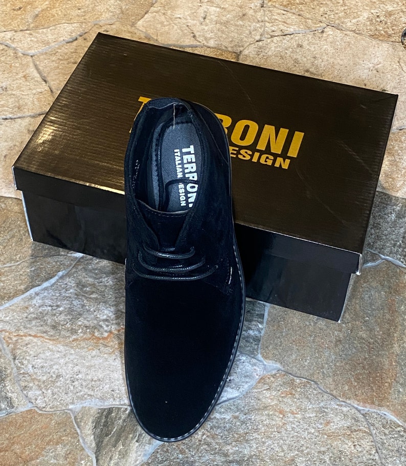 May include: Black suede chukka boot with a lace-up design, displayed next to a black box. The box features gold lettering that reads "TERRONI DESIGN". The shoe has a stitched sole and a soft, textured appearance.