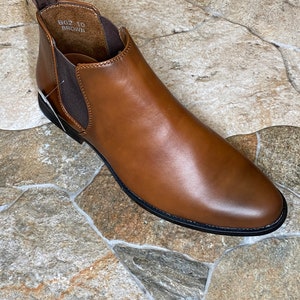 May include: A brown leather Chelsea boot with a rounded toe and elastic side panels. The boot has a pull tab at the back and a black sole. The text "B02 10 BROWN" is visible inside the boot.