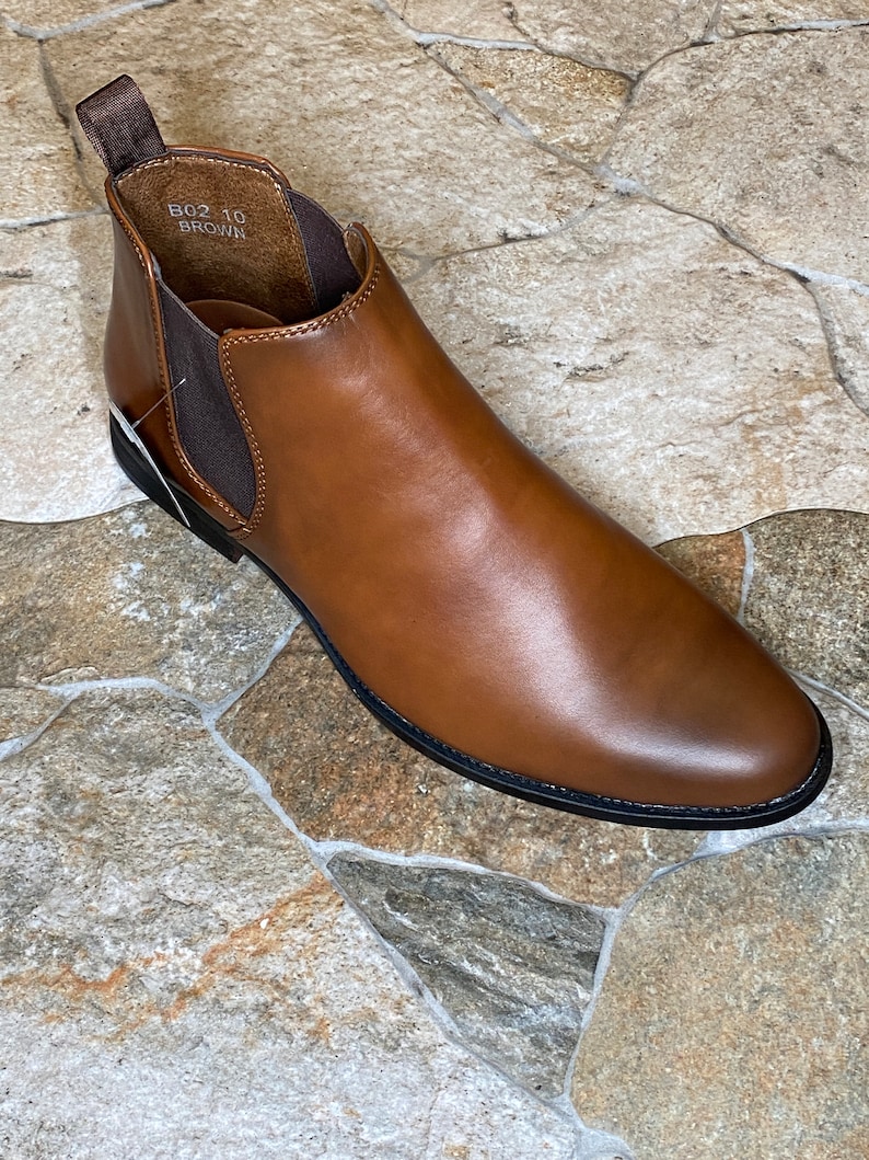 May include: A brown leather Chelsea boot with a rounded toe and elastic side panels. The boot has a pull tab at the back and a black sole. The text "B02 10 BROWN" is visible inside the boot.