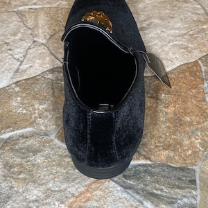 May include: Black velvet loafer with a gold-coloured decorative accent. The shoe has a rounded toe and a low heel. A tag is attached to the side. The shoe is designed for formal occasions.