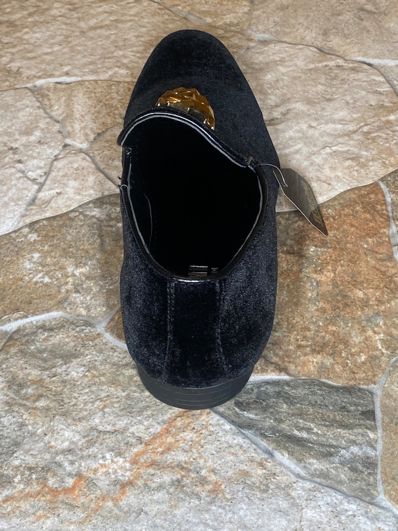 May include: Black velvet loafer with a gold-coloured decorative accent. The shoe has a rounded toe and a low heel. A tag is attached to the side. The shoe is designed for formal occasions.