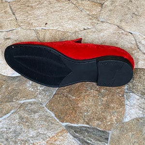 May include: A red velvet loafer with a black sole. The shoe's sole has a textured design for grip. The shoe is set against a stone tile background.