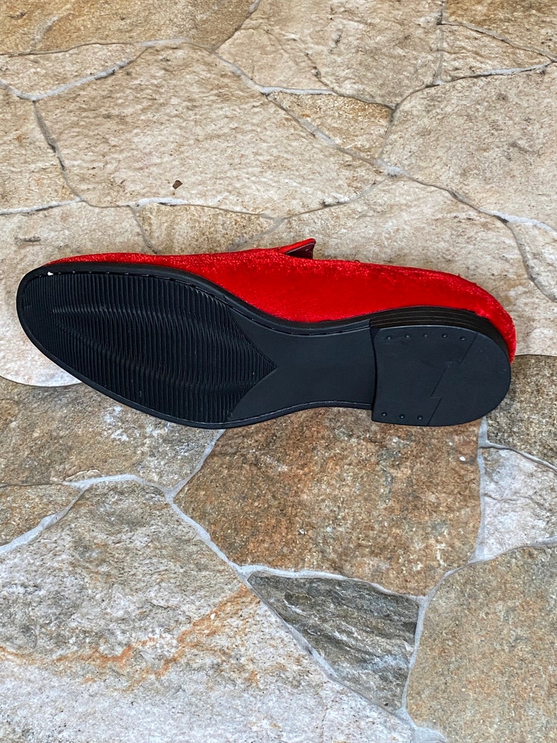 May include: A red velvet loafer with a black sole. The shoe's sole has a textured design for grip. The shoe is set against a stone tile background.