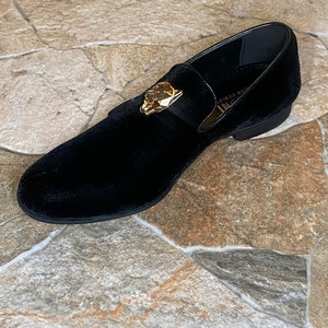 May include: Black velvet loafer with a gold-coloured decorative accent. The shoe has a rounded toe and a low heel. The shoe is on a stone surface.