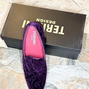 Op de afbeelding: Een paar paarse fluwelen loafers met een sierlijk bloemmotief. De schoenen hebben een roze binnenkant en liggen naast een zwarte doos met de witte tekst "TERRONI DESIGN".