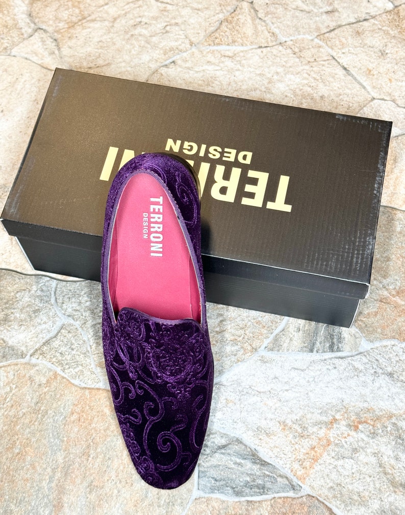 Op de afbeelding: Een paar paarse fluwelen loafers met een sierlijk bloemmotief. De schoenen hebben een roze binnenkant en liggen naast een zwarte doos met de witte tekst "TERRONI DESIGN".