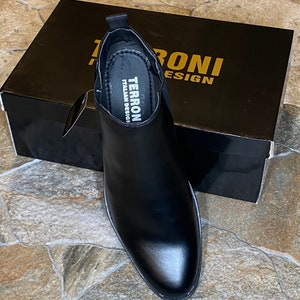 Puede incluir: Bota Chelsea de cuero negro con punta afilada, frente a una caja negra. La caja tiene letras doradas que dicen "TERRONI DESIGN". La plantilla de la bota dice "TERRONI ITALIAN DESIGN".