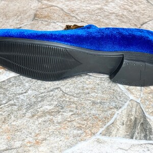 May include: A close-up of a blue velvet loafer with a black sole. The shoe features a gold-colored buckle and a textured sole. The shoe is approximately 11 inches long.